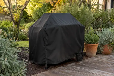 Housse noire en tissu Oxford couvrant un barbecue dans un jardin avec terrasse en bois et feuillage.